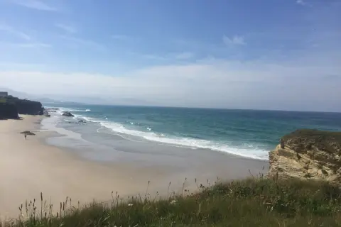 Un paseo solitario por las **playas de Lugo**, con el mar turquesa rompiendo suavemente en la arena dorada y acantilados rocosos.