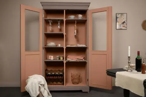Une vieille armoire repeinte, aux portes roses et intérieur gris, abrite vaisselle, verres à pied, bouteilles de vin et paniers.