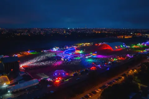 Nocne, kolorowe światła rozświetlają teren festiwalu na powietrzu. Tłumy bawią się przy scenach i namiotach, a w tle widać miasto.