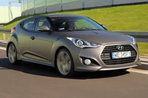 Szary Hyundai Veloster pędzi po drodze, za nim zielone ekrany dźwiękochłonne.