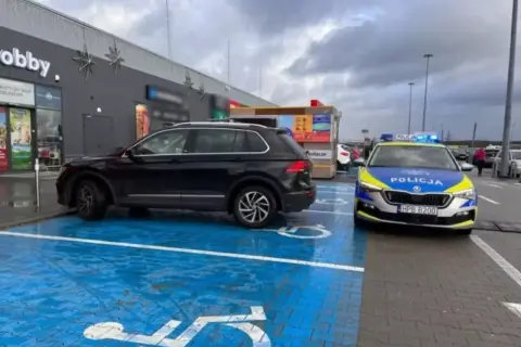 Czarny SUV zaparkowany na niebieskim miejscu dla niepełnosprawnych, obok radiowóz policyjny. To pytanie o to, jak długo można parkować na miejscu dla niepełnosprawnych, jest tu bardzo aktualne.