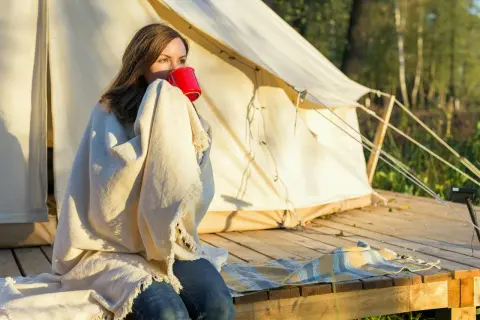 Kvinna med filt och röd mugg vid tältet. En mysig morgon som visar vikten av en bra packlista camping.