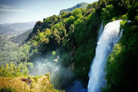 Cascata imponente nella rigogliosa montagna umbria, con valli verdi e cielo azzurro.