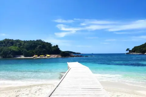 Drewniany pomost prowadzi do krystalicznie czystej wody na **Ksamil beach**. W oddali widać zielone wyspy i żółte łódki.