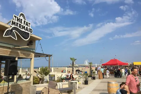 Strandpromenade in Den Haag mit dem Restaurant "The Fat Mermaid", Menschen, Sonnenschirmen und einem Riesenrad. Tolle Den Haag Unternehmungen!