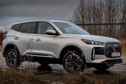 Srebrny Chery Tiggo 4 na tle ponurego krajobrazu. Auto stoi na mokrej nawierzchni, otoczone suchymi trawami i drzewami.