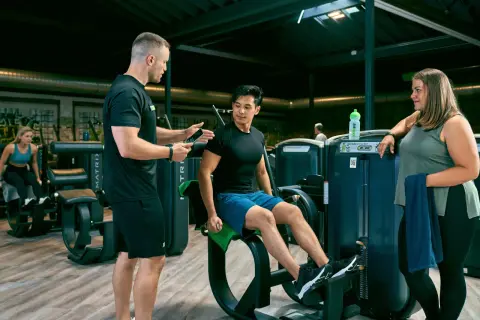Trainer erklärt Mann die richtige Ausführung am Gerät für den erector spinae. Frau schaut zu.