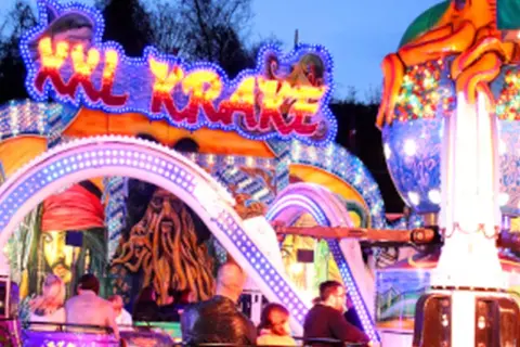 Bunte Lichter und Piratenmotive auf einer Kirmes in Berlin. Eine Achterbahn mit dem Schriftzug "Sky Krake" zieht Besucher an.