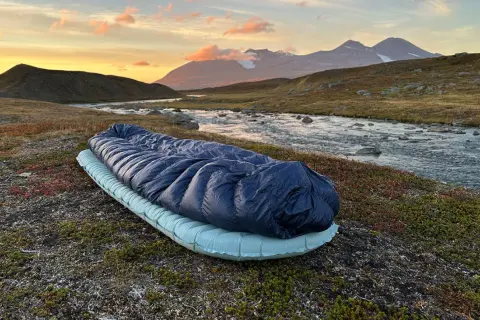 Sovsäck och liggunderlag redo för en natt i fjällen. Test av liggunderlag vid en forsande älv i solnedgången.