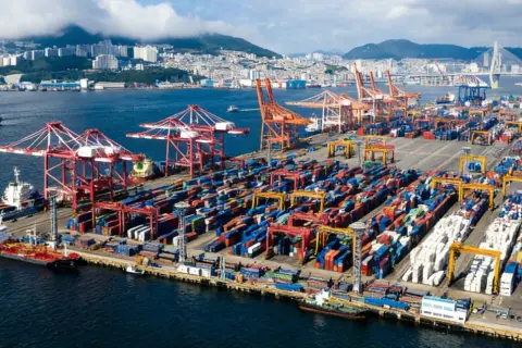 Ogromny port kontenerowy z dźwigami i statkami. W tle widać miasto i most. To skrót do globalnego handlu.