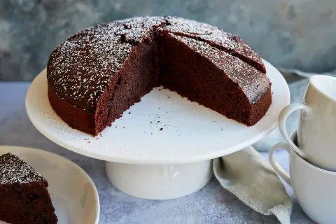 Ein saftiger veganer Schokokuchen, bestäubt mit Puderzucker, steht auf einem weißen Tortenständer. Daneben liegen Tassen und eine Serviette.