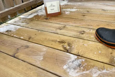 En flaska linoljesåpa står på en altan som håller på att rengöras. En svart stövel med orange kant syns i nedre högra hörnet.