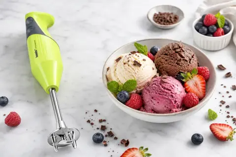 Zielony blender Bamix obok miski z lodami waniliowymi, czekoladowymi i malinowymi, ozdobionymi owocami. Idealny do domowego sposobu, jak zrobić lody.