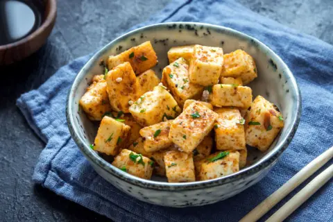 Usmażone kostki tofu, które jest zrobione z ziaren soi, w misce z sezamem i ziołami. Obok pałeczki i sos sojowy.