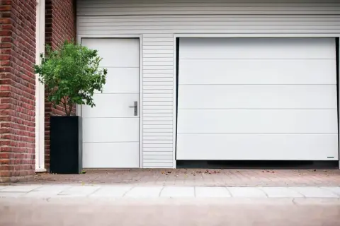 Modernt vipport garage med en vit port och en sidodörr. En grön buske i en svart kruka står bredvid.