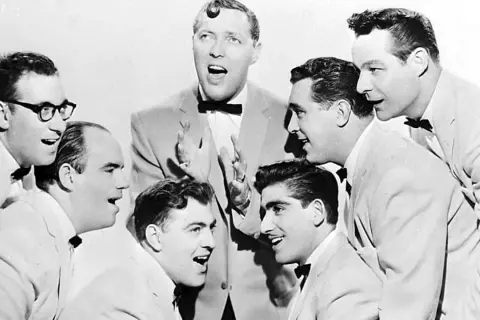 Gruppe von Männern in Anzügen und mit Fliegen, die singen.