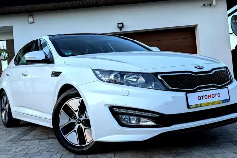 Biały kia sedan z czarnym dachem, zaparkowany przed domem z drewnianą bramą garażową.