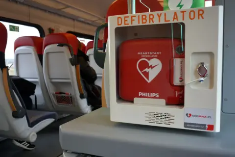 Czerwony defibrylator AED w białej obudowie, zamontowany w pociągu, obok siedzeń.