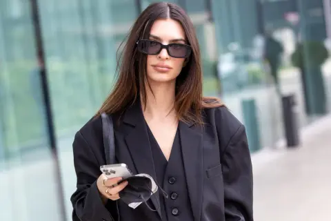 Elegancka brunetka w okularach przeciwsłonecznych, z telefonem i maseczką w dłoni. Jej włosy mają piękny, ciemny kolor.