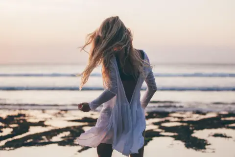 Blondynka w zwiewnej białej sukience tańczy na plaży, a wiatr rozwiewa jej włosy. W tle widać spokojne beach waves i zachodzące słońce.