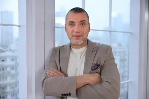 Maciej Dębosz prywatnie, w beżowej marynarce i z dumnym spojrzeniem, opiera się o okno.
