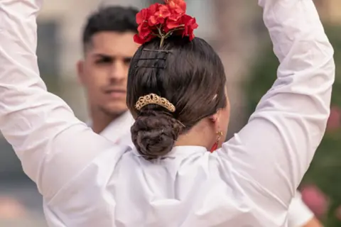 Peinados para bailar flamenco: moño bajo adornado con flores rojas y peineta dorada. Un hombre con camisa blanca al fondo.