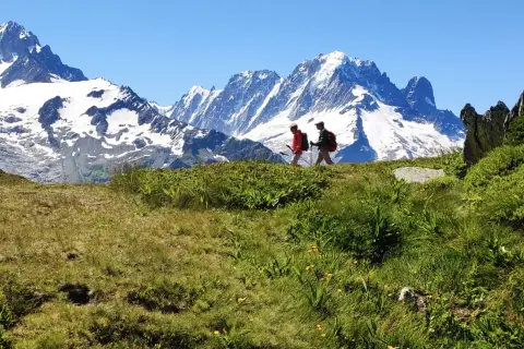Deux randonneurs marchent sur une crête herbeuse face à des montagnes enneigées.