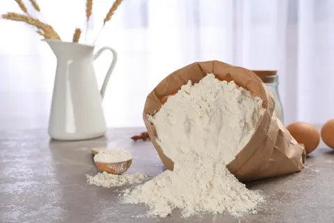 Wsypana na blat mąka ze zboża, obok drewniana łyżka, dzbanek ze snopkami zbóż i jajka.