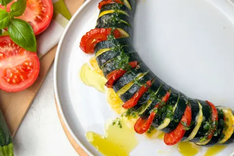 Zucchini Caprese: geschichtete Zucchini, Tomaten und Kräuter, beträufelt mit Olivenöl auf einem weißen Teller.