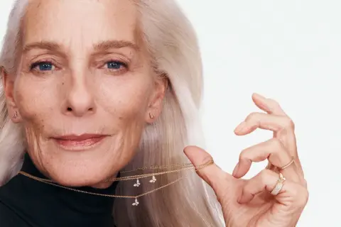 Mujer mayor con cabello plateado, luciendo una medalla de la madre con forma de mariposa y anillos brillantes.
