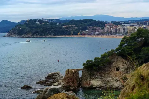 Widok na zatokę z łodziami i plażą. W oddali widać miasto i góry. Pogoda na Costa Brava zapowiada się na spokojną.