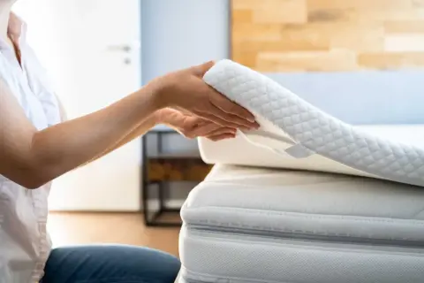 Ręce układają biały, pikowany topper na łóżko, poprawiając komfort snu.