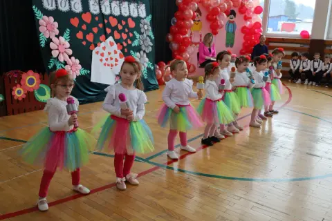 Dzieci w kolorowych spódniczkach śpiewają piosenkę na Dzień Dziecka, trzymając w rękach mikrofony.