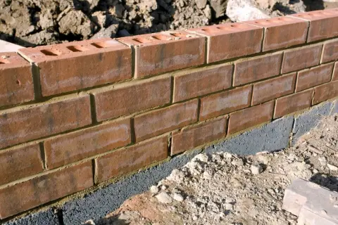 Ceglana ściana ogrodzenia z widocznym szarym bloczkiem fundamentowym, który stanowi alternatywę dla tradycyjnej podmurówki.