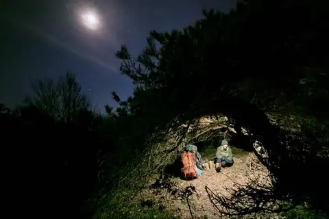 Två personer sitter under en grenig trädkrona, redo att sova under bar himmel. En ryggsäck står bredvid. Månen lyser starkt på natthimlen.