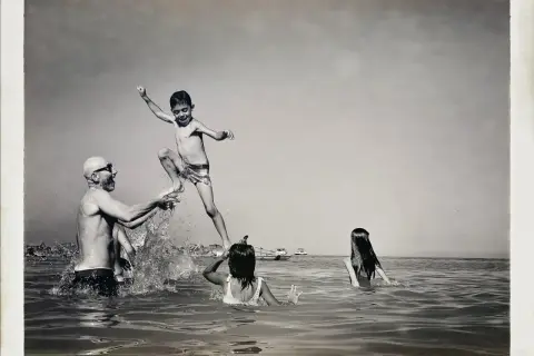 Un bambino salta in aria, sorretto da un uomo con occhiali da sole, mentre due bambine nuotano. Un momento di gioia estiva, un ricordo di Carlo Traini.