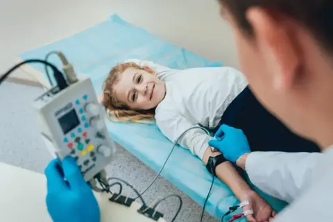 Uśmiechnięta dziewczynka leży na łóżku podczas badania EEG, które pomaga diagnozować padaczkę u dzieci.