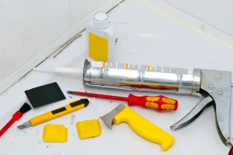 Werkzeuge zum Silikon von Fliesen entfernen: Kartuschenpresse, Cutter, Schaber, Bürste und eine Flasche mit Reiniger.