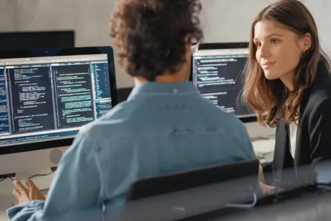 Dwóch programistów przy komputerach, jeden w pomarańczowej czapce, drugi w dżinsowej koszuli. Rozmawiają o tym, jak się przebranżowić na IT.