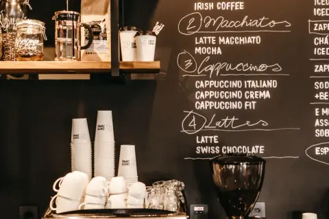 Tafel mit Kaffeebar Ideen: Macchiatos, Cappuccinos, Lattes und mehr. Stapel von Pappbechern und Espressomaschine im Vordergrund.