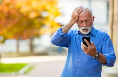 Anziano con mano sulla testa, guarda il telefono con espressione preoccupata, forse per la paura di parlare al telefono.