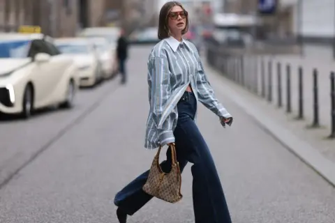 Kobieta w luźnej, pasiastym koszuli i dżinsach pokazuje, jak nosić koszule w miejskim stylu. Torba na ramię dodaje elegancji.