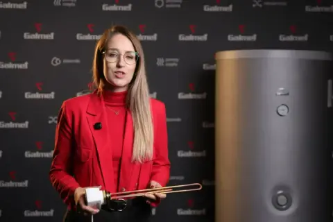 Kobieta w czerwonej marynarce prezentuje element grzałki elektrycznej, kluczowy dla sprawności grzałki elektrycznej. Obok stoi bojler.