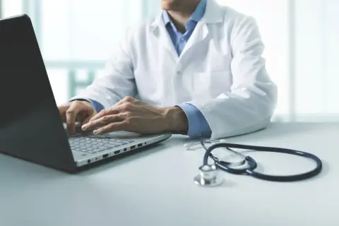 Lekarz w białym kitlu wykonuje badania wstępne do pracy, wpisując dane na laptopie. Obok leży stetoskop.