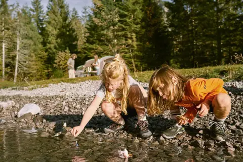 Två barn leker med pappersbåtar vid en sjö. I bakgrunden syns ett tält och träd. En härlig 5 kamp fest i naturen.