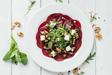 Rote Bete Carpaccio mit Rucola, Walnüssen und Feta auf einem weißen Teller, garniert mit frischem Basilikum.