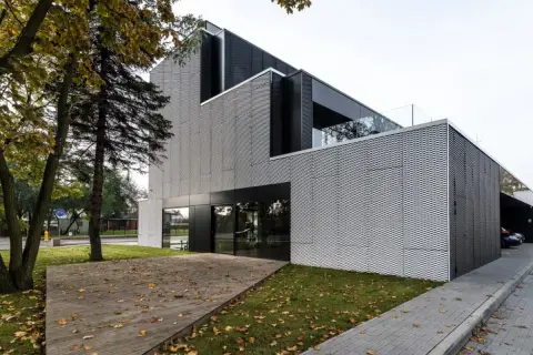 Nowoczesny budynek z metalicznym ryzalitem, przeszklonym wejściem i drewnianym tarasem porośniętym jesiennymi liśćmi.