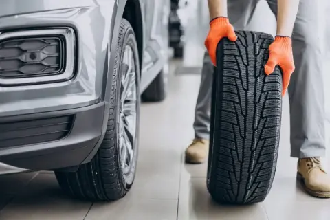 Mechanik w pomarańczowych rękawicach trzyma nowe opony asymetryczne obok szarego samochodu.