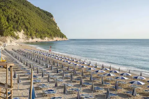 Filettata di ombrelloni e lettini sulla spiaggia di San Michele, con il mare azzurro e la collina verde sullo sfondo.