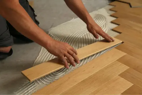 Manos colocando tablones de maderas duras sobre adhesivo para crear un suelo de espiga.